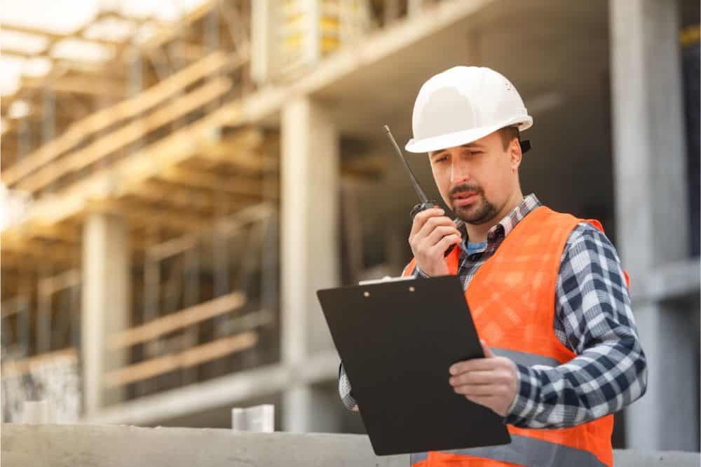 pxton _PX-888S Walkie Talkies Review engineer wearing white safety vest and hardhat with walkie talkie and clipboard inspecting construction site